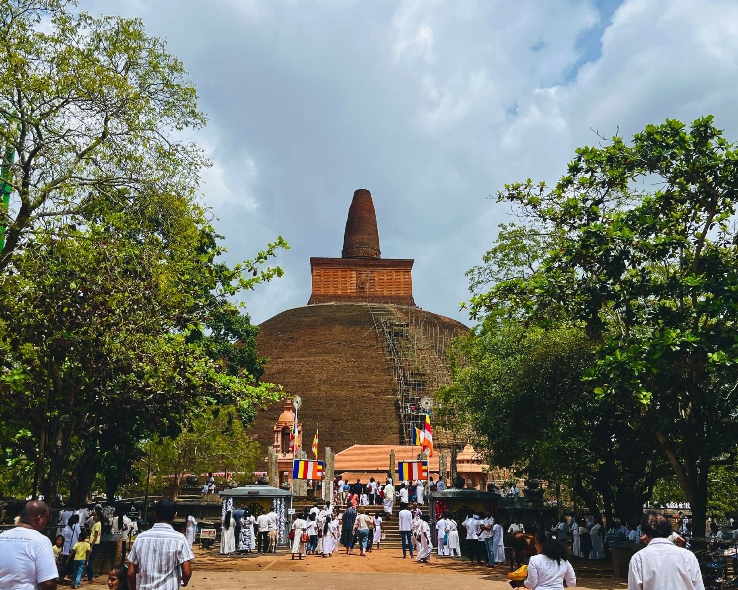 Abhayagiri Stupa massive exposed brick dome with harmika and spire framed by trees, pilgrims in white, Buddhist flags and scaffolding at main entrance in Anuradhapura