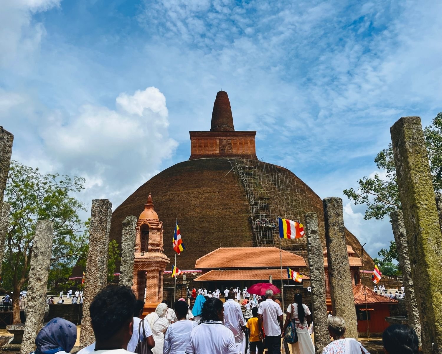 Abhayagiri Stupa massive brick dome with spire seen through ancient stone pillars at south entrance, pilgrims in white walking toward orange-tiled wahalkada under blue sky in Anuradhapura