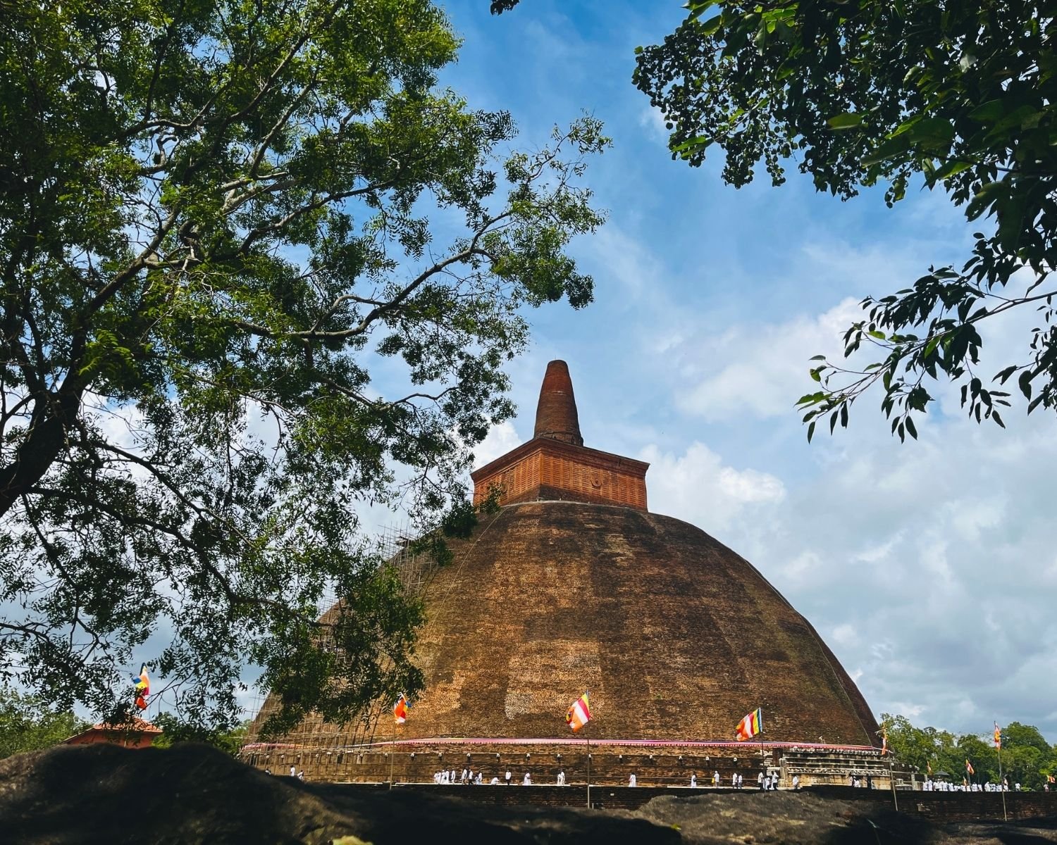 Abhayagiri Stupa massive ancient brick dome seen from southeast through trees, pilgrims on stone platform at base reveal the immense 75m height, bamboo scaffolding left in Anuradhapura