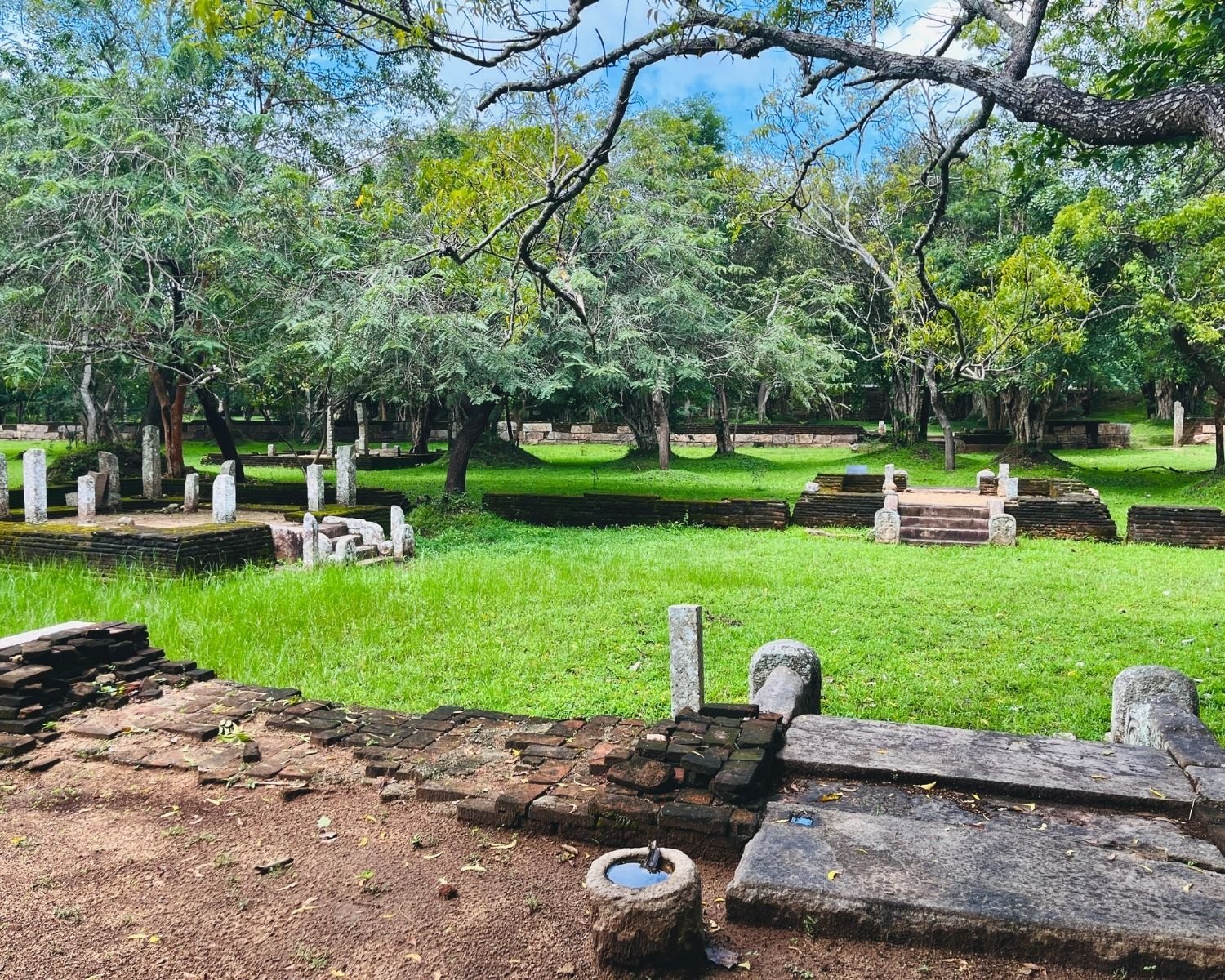 Multiple stone platforms with steps and pillars across grassy courtyards with a stone water basin in the residential complex at Abhayagiri in Anuradhapura