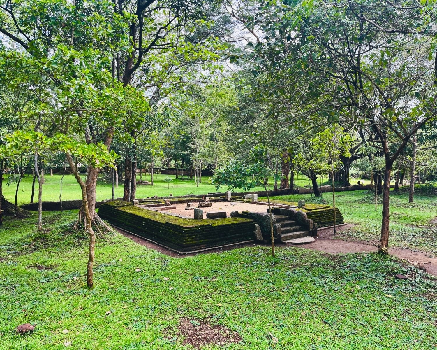 Small moss-covered stone shrine with steps and guardstones sits among trees near Abhayagiri Banyan Tree Shrine in Anuradhapura