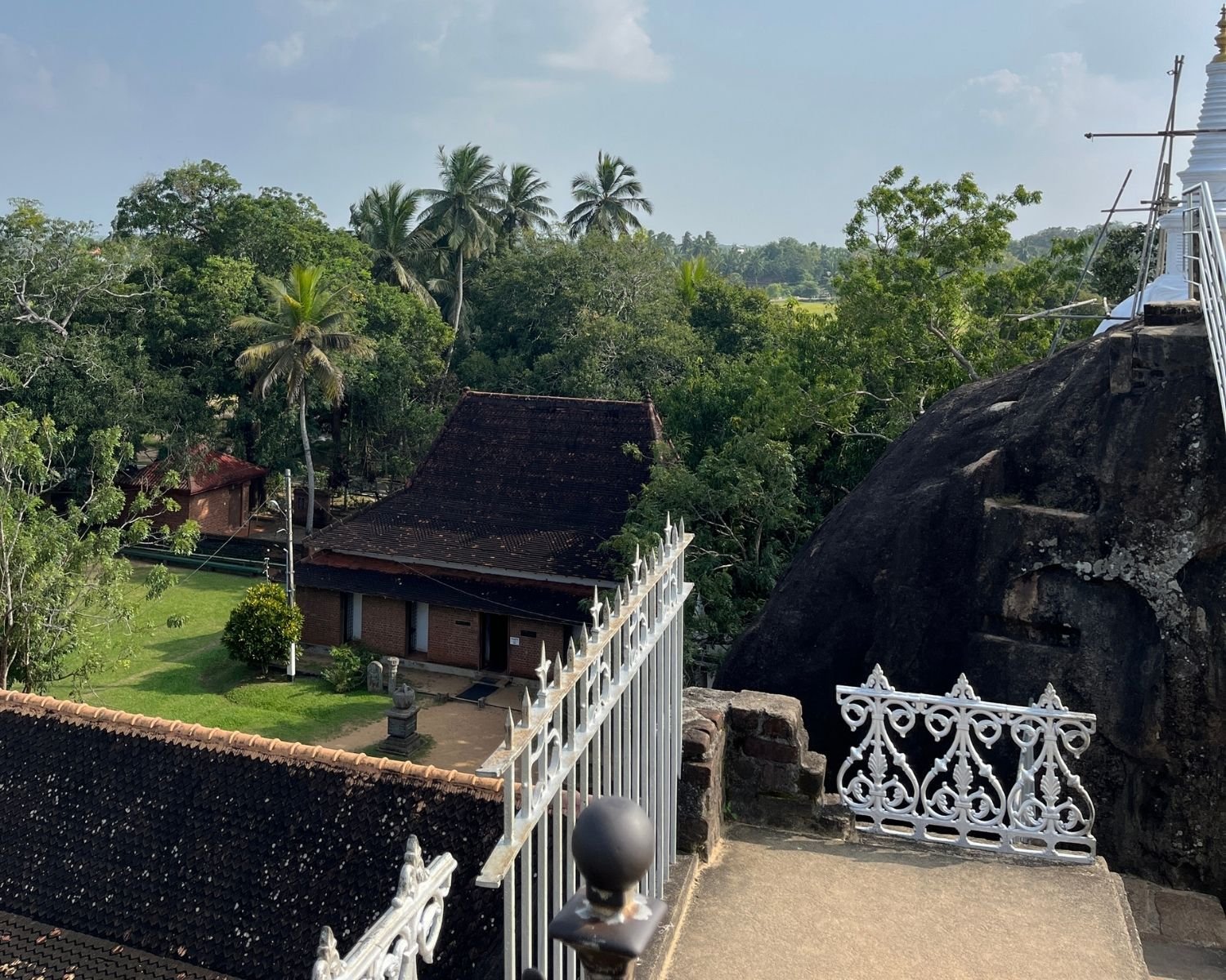 Carved rock niches in black boulder at Isurumuniya temple, with ornate white iron railings, brick shrine with dark tiled roof, manicured lawn and coconut palms under blue sky at Anuradhapura