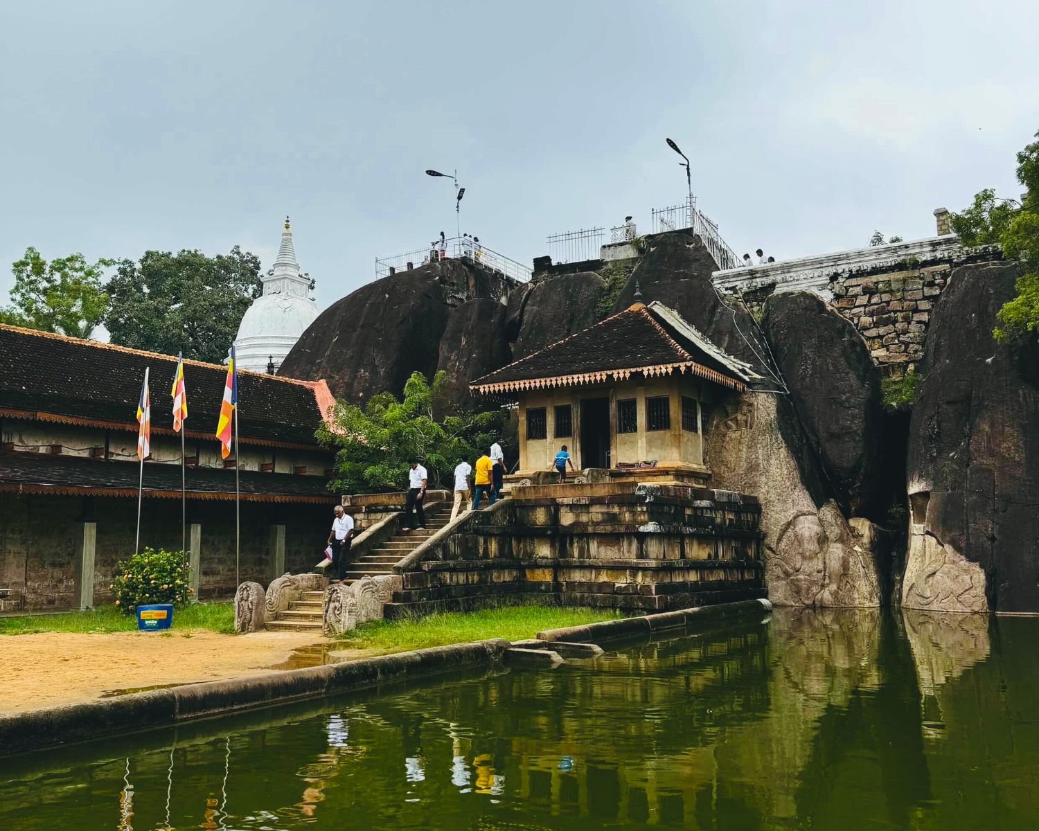 Isurumuniya rock temple in Anuradhapura with ancient cave shrine on stone platform, granite outcrop, Buddhist flags, lotus pond reflection, white stupa, and pilgrims climbing steps
