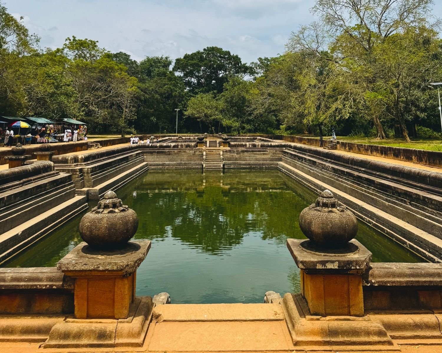 Kuttam Pokuna twin pools seen head-on with carved punkala urns on granite balustrades, tiered steps descending to reflective green water, market stalls with colorful umbrellas and trees ringing the site at Anuradhapura