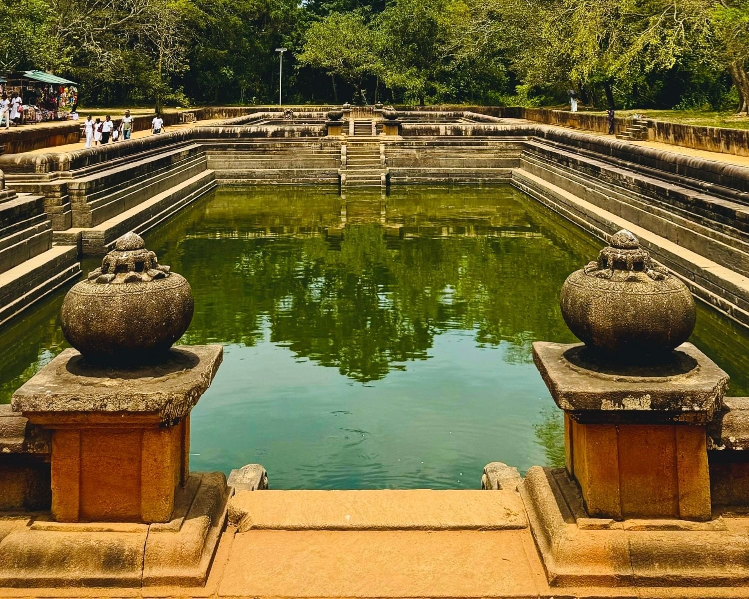 Carved punkala urns frame the reflecting green pools of Kuttam Pokuna, with granite tiers and steps leading to mirrored tree canopy, market stalls and forest backdrop at Anuradhapura