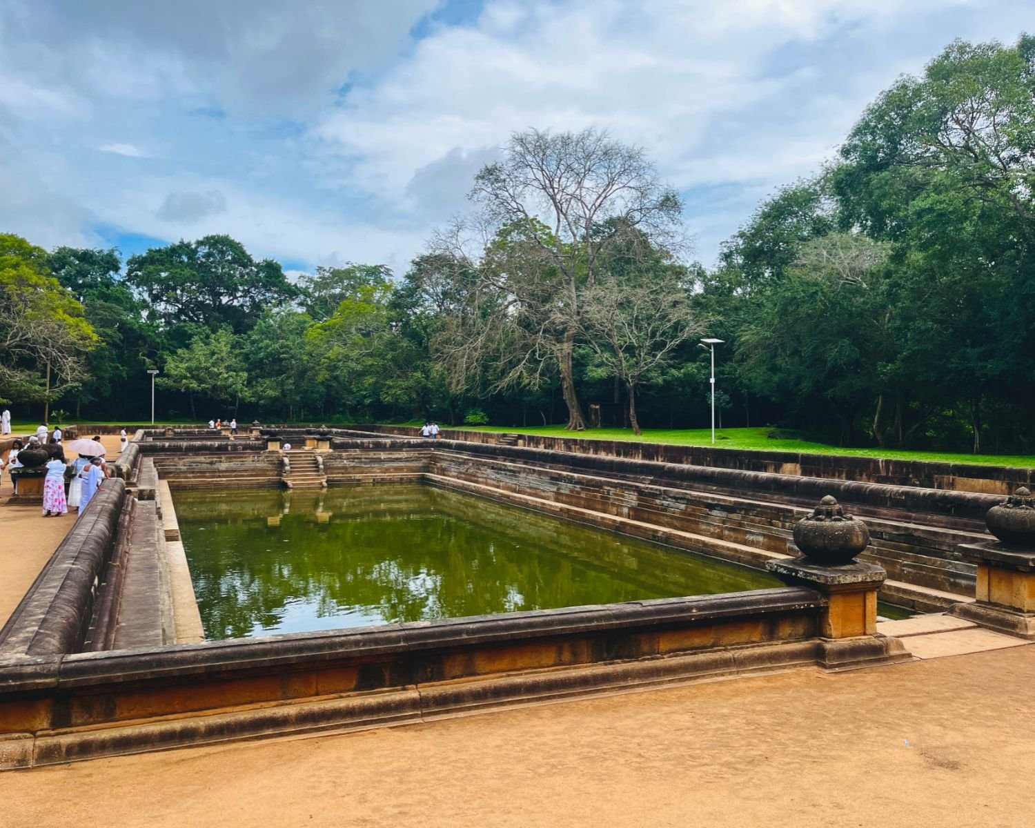 Kuttam Pokuna twin pools stretch under blue sky with scattered clouds, lined by carved punkala urns on granite tiers, with leafless tree and green canopy beyond at Anuradhapura