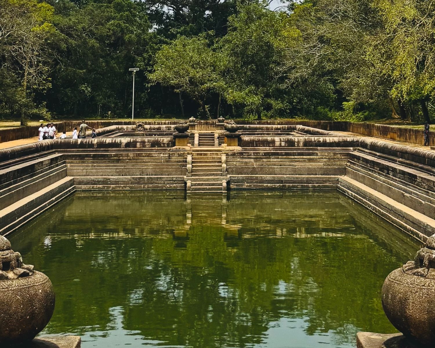 Kuttam Pokuna twin bathing pools with carved stone punkala urns in foreground, granite steps descending to green water, pilgrims in white and dense forest canopy beyond at Anuradhapura