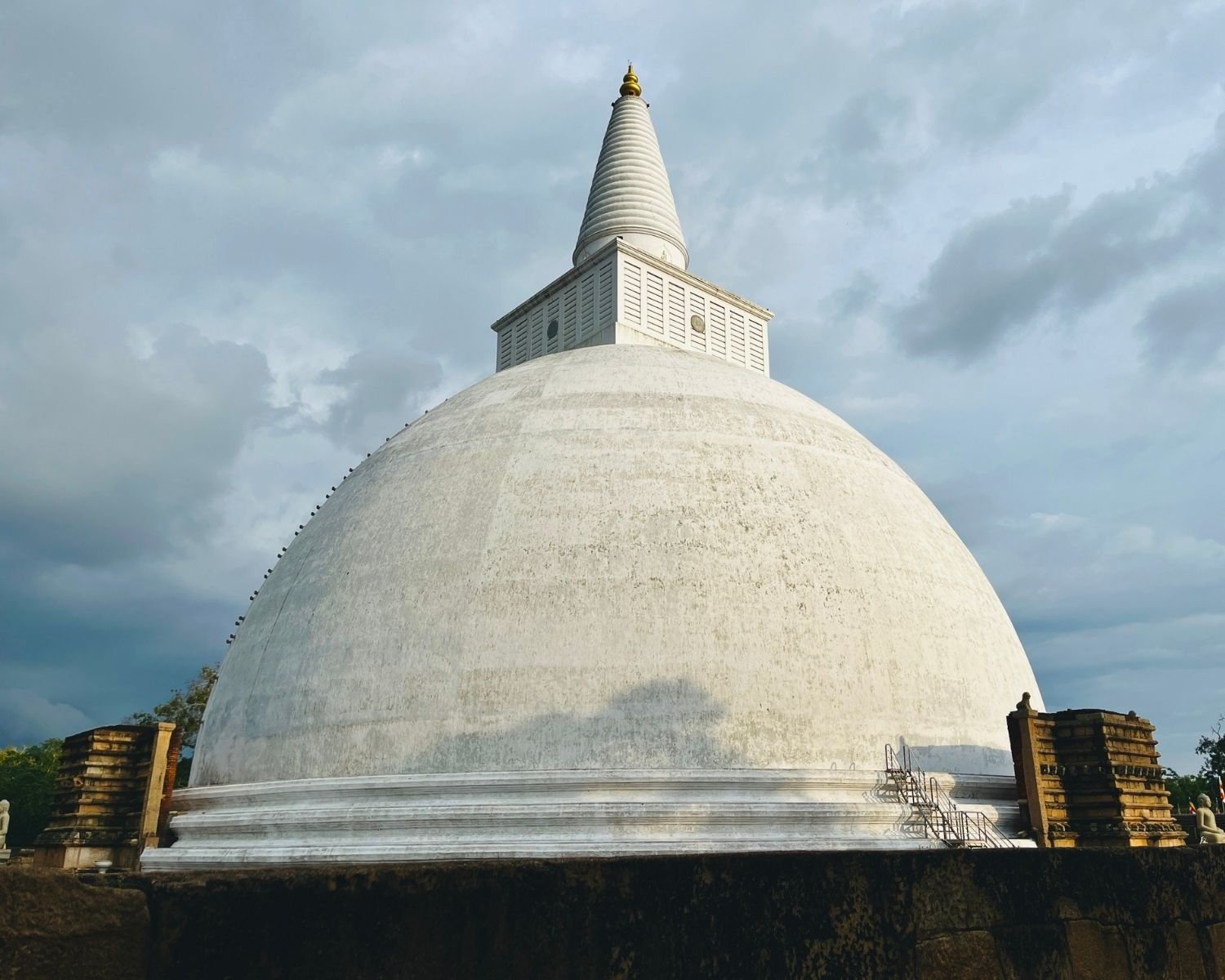 Mirisawetiya Stupa white dome at northwest with twin ancient brick vahaalkada ruins, cloud shadow on dome, stone sailcloth pegs, stone platform and meditating Buddha statue under cloudy sky at Anuradhapura