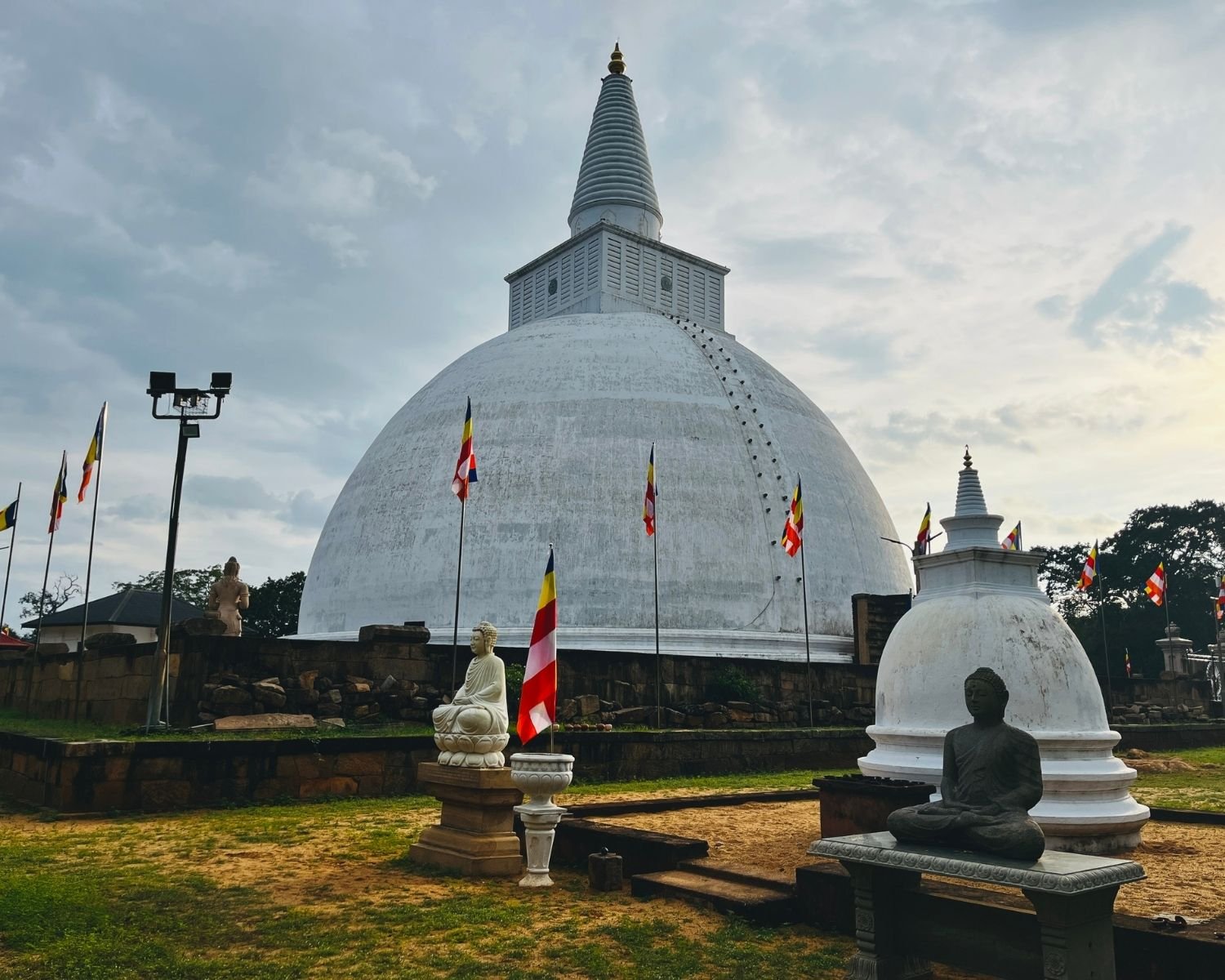 Mirisawetiya Stupa white dome towers behind small stupa with Buddha statues in meditation and standing poses, Buddhist flags, stone platform and grass courtyard under cloudy sky at Anuradhapura