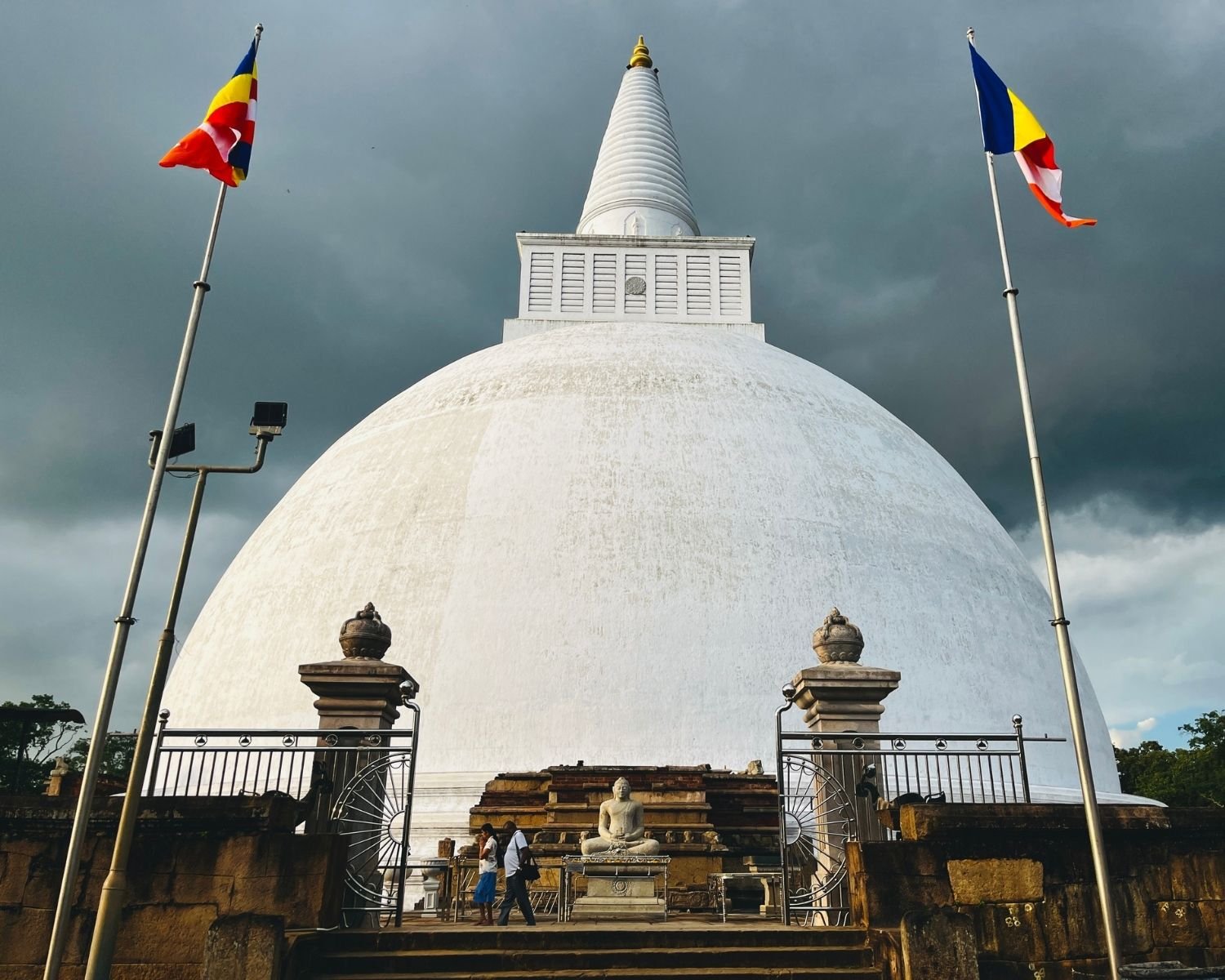 Mirisawetiya Stupa white dome sunlit after drizzle at south entrance, with Buddhist flags, ornate gate with wheel motifs, Buddha statue in meditation, stone platform and bright blue sky at Anuradhapura