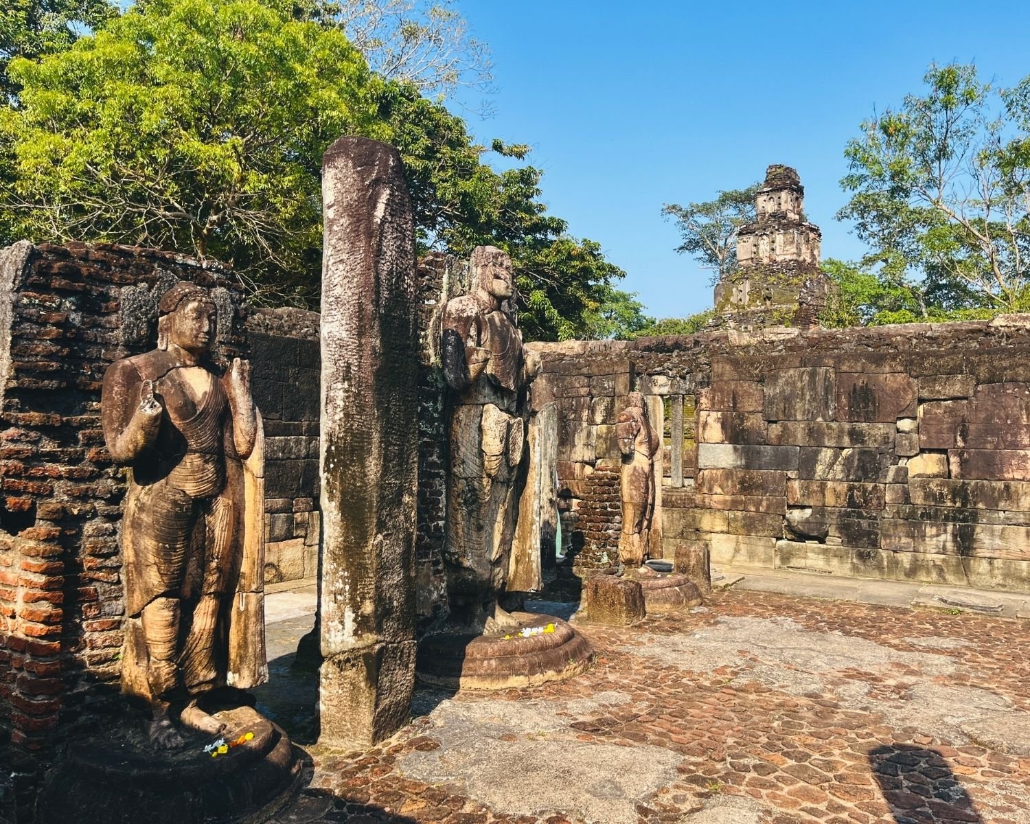 Three standing Buddha statues in Hatadage ruins with stone pillars and Satmahal Prasada tower behind