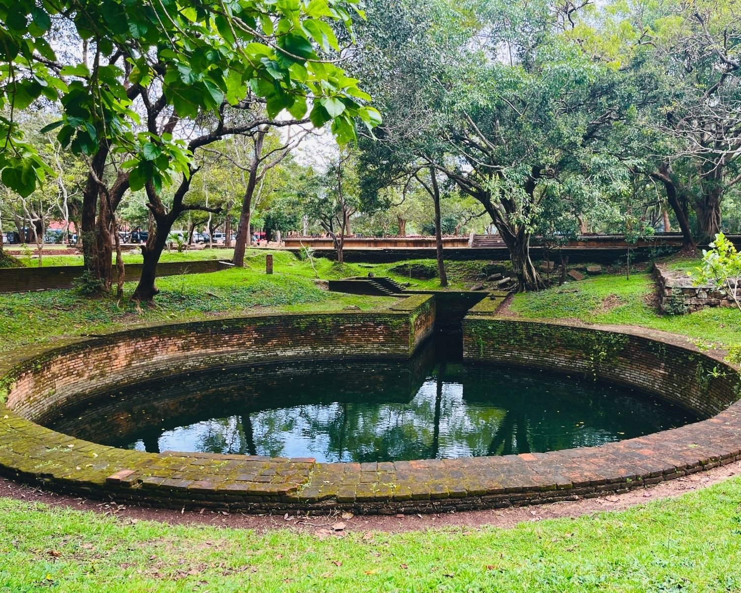 Royal Key Cistern brick basin with straight inlet channel reflecting trees, surrounded by ancient green grounds, mossy walls, stone steps, and large shade trees at Abhayagiri monastery Anuradhapura