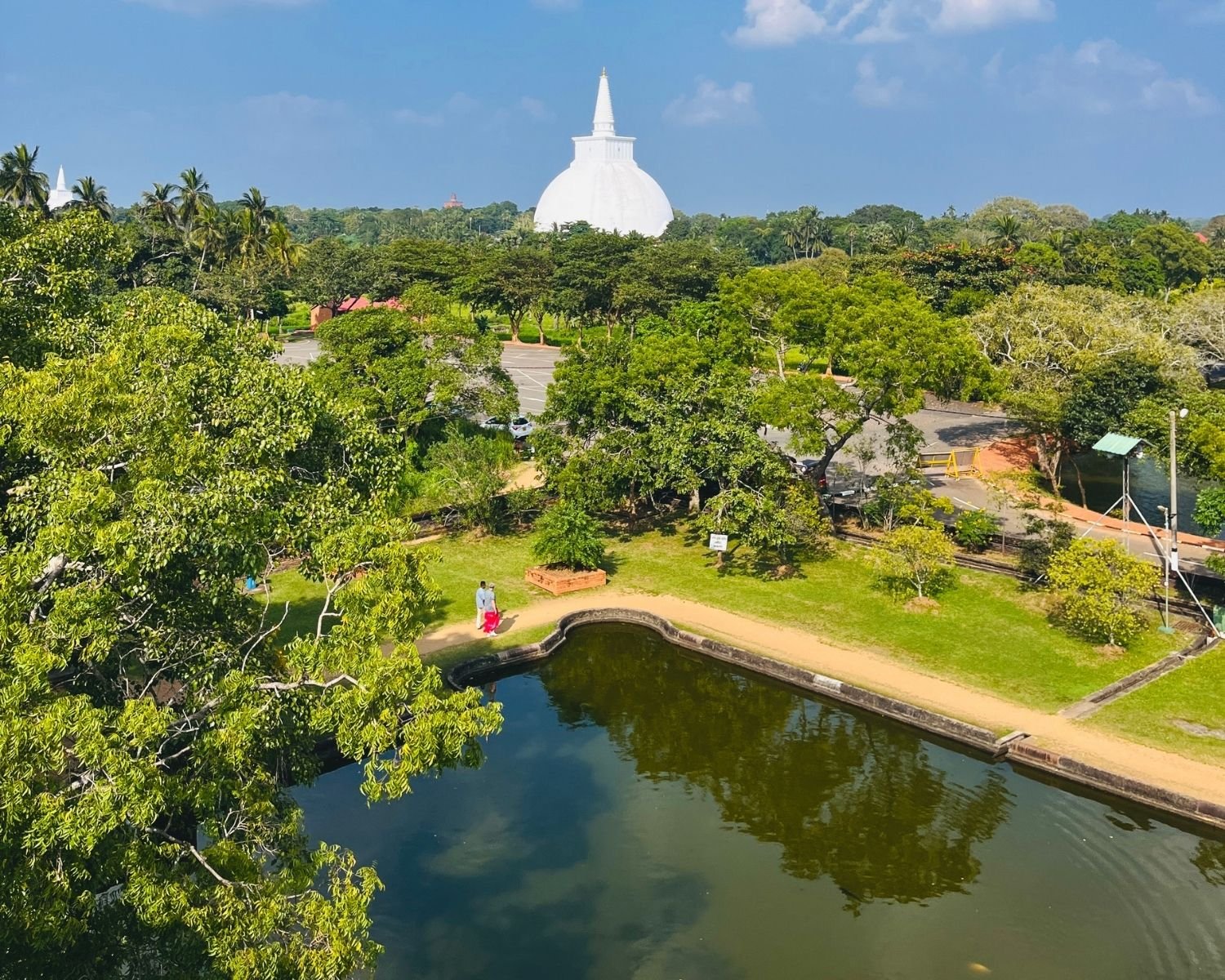 White dome of Sanda Hiru Seya stupa rising beyond trees and parkland, with Isurumuniya Temple Pond, dirt path, green lawn and coconut palms under blue sky at Anuradhapura