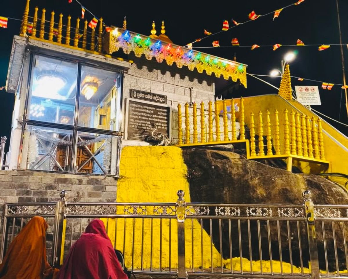 Sri Pada sacred footprint shrine at Adams Peak summit at night with gold railing colorful lights Buddhist flags and pilgrims praying before the yellow painted rock