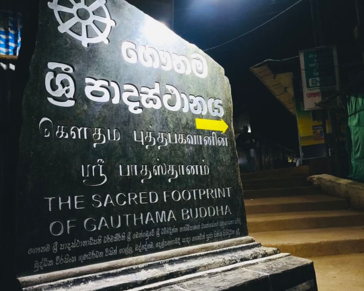Stone trailhead sign at Nallathanniya for Sri Pada Adams Peak at night with trilingual text Sacred Footprint of Gauthama Buddha and yellow arrow pointing up steps