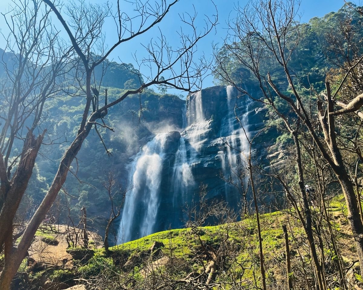 Bomburuella Waterfall full drop seen from hiking trail with sunlit grass and bare trees framing misty cascades in Nuwara Eliya Sri Lanka