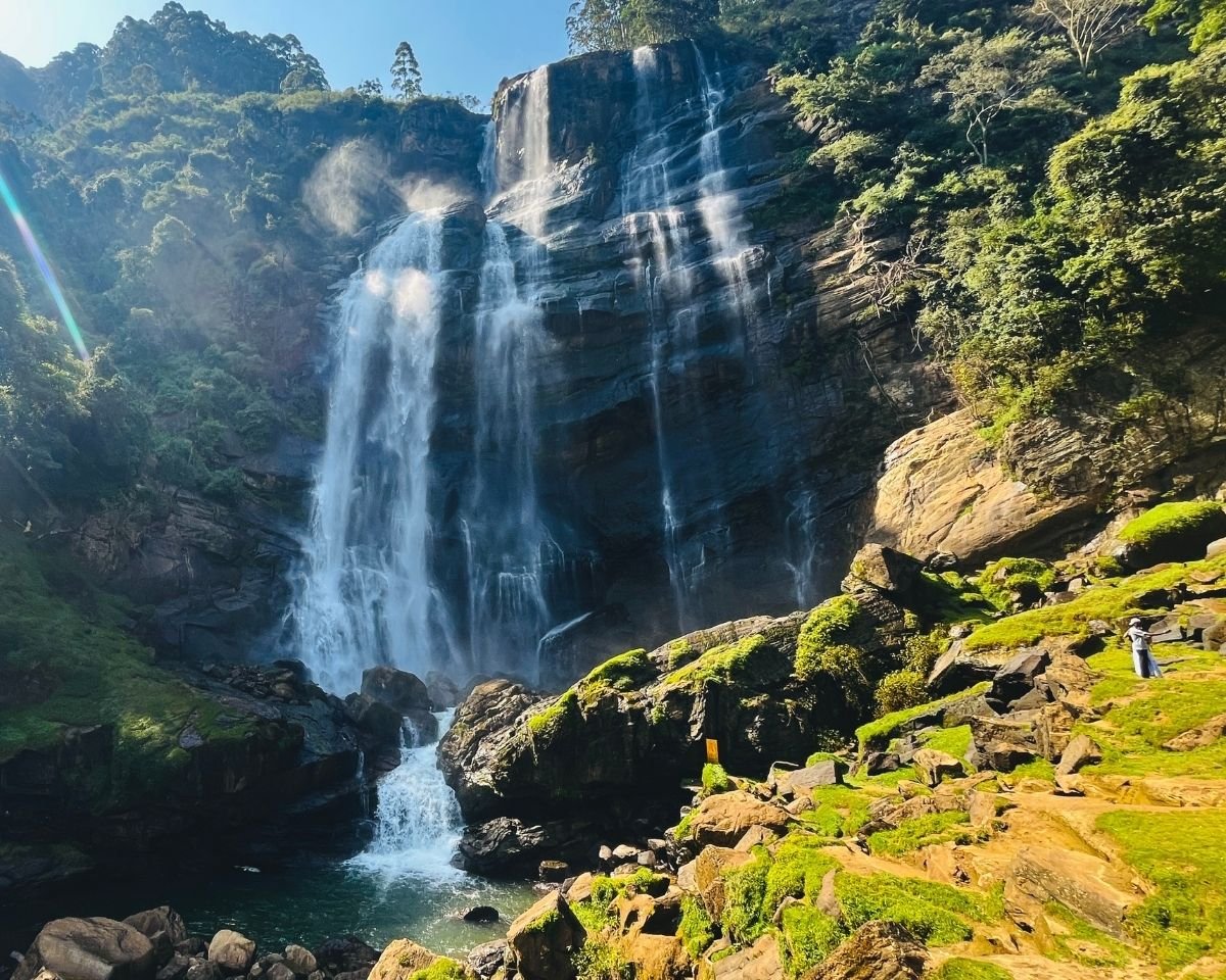 Bomburuella Waterfall sunlit cascades flowing down wide rock face surrounded by vibrant green forest and mist in Nuwara Eliya Sri Lanka