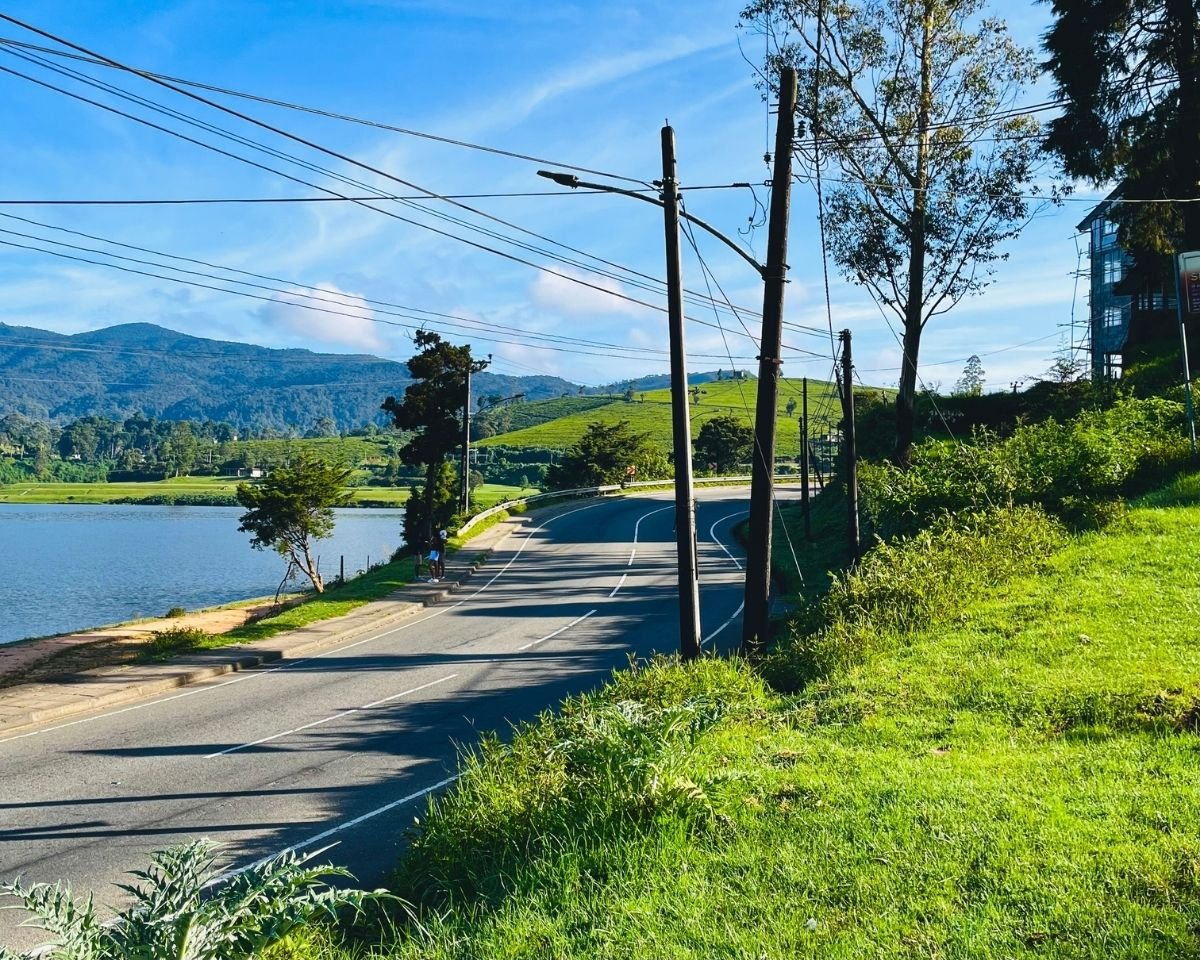 Gregory Lake with lakeside road, power lines, tea estate hills, and mountains under blue sky Nuwara Eliya