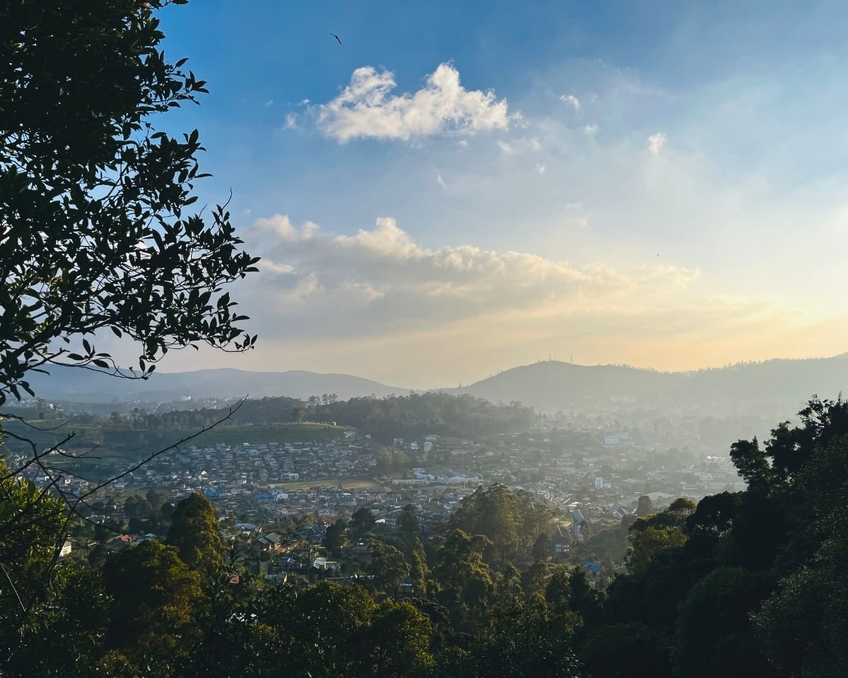 View of Nuwara Eliya town and Single Tree Hill from the base of Lovers Leap Falls framed by trees under blue sky Sri Lanka