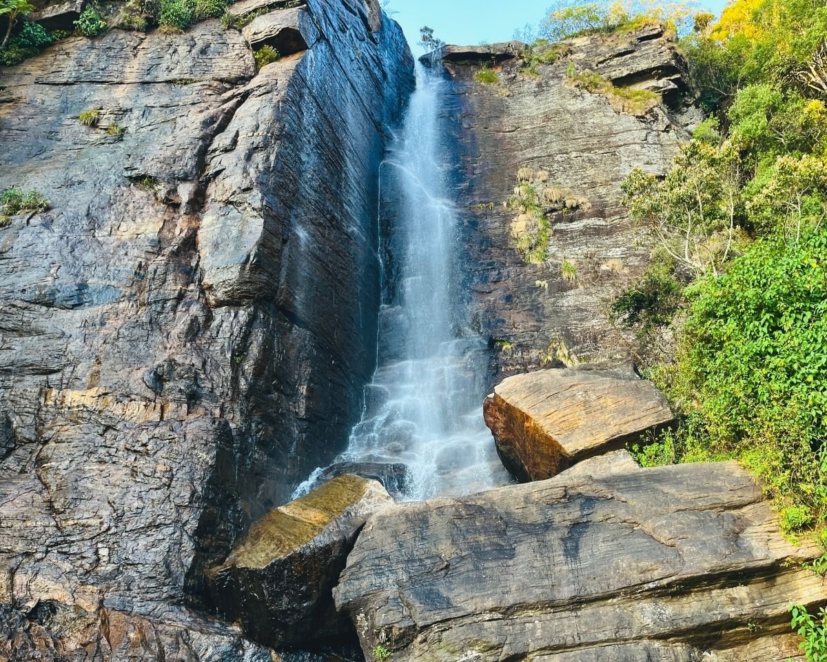 Lovers Leap Falls waterfall flowing between dark rock cliffs with boulders and green trees under blue sky Nuwara Eliya Sri Lanka
