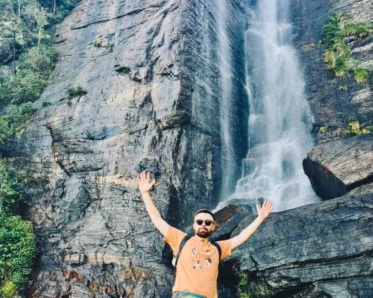 Person with raised arms at base of Lovers Leap Falls waterfall with rock cliffs and forest Nuwara Eliya Sri Lanka