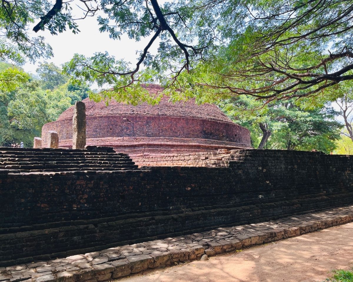 Ancient brick stupa dome behind dark weathered brick outer wall with stone pillars and green trees at Dambulla Somawathiya archaeological site Sri Lanka