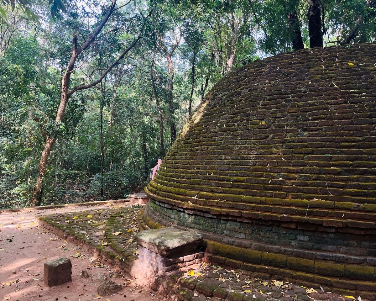 Side view of ancient mossy brick stupa with hiker in background inside Jathika Namal Uyana ironwood forest Dambulla Sri Lanka